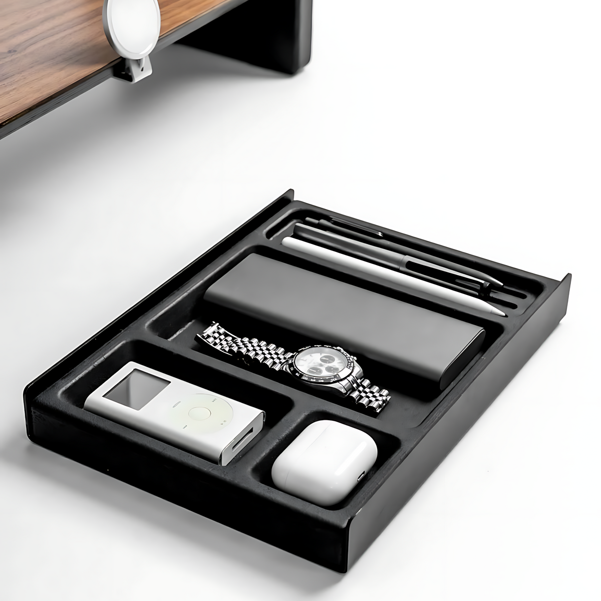 A black desk organizer tray with multiple compartments holding a white iPod, white earbud case, pen, and a watch, shown on a clean white surface.